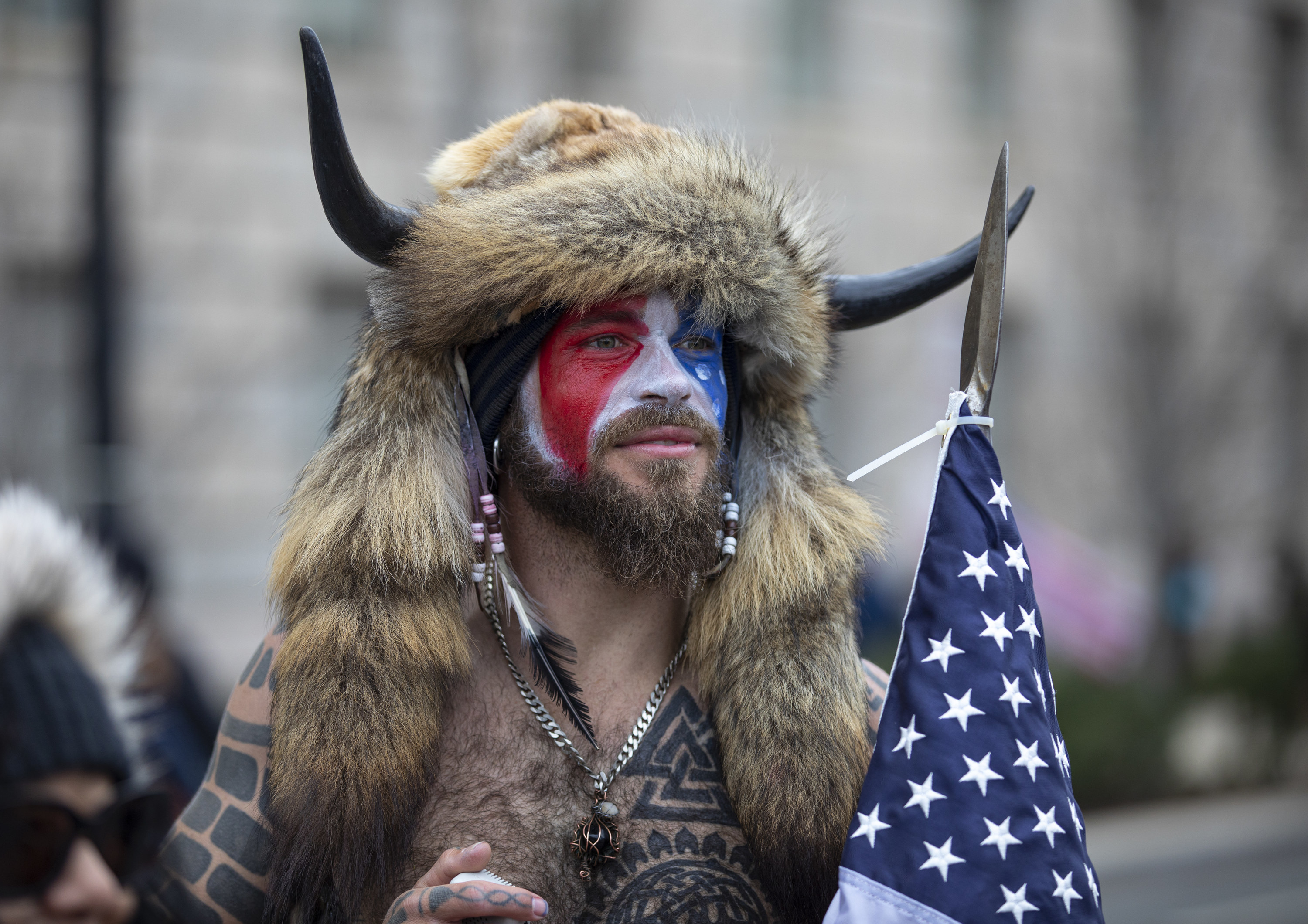 Chansley wearing a fur hat with horns and carrying an American flag with red, white, and blue face paint on