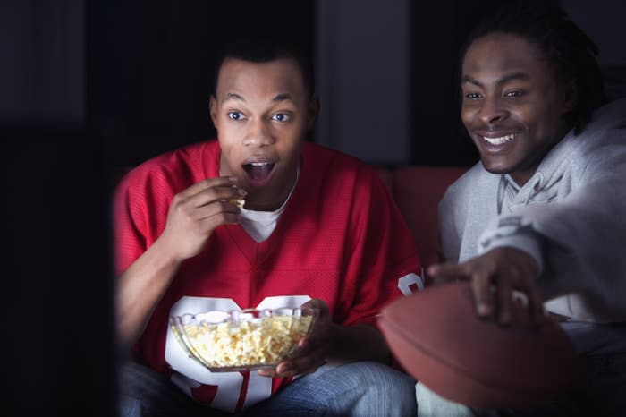 Two men in football entire watching the TV while eating popcorn and holding a football
