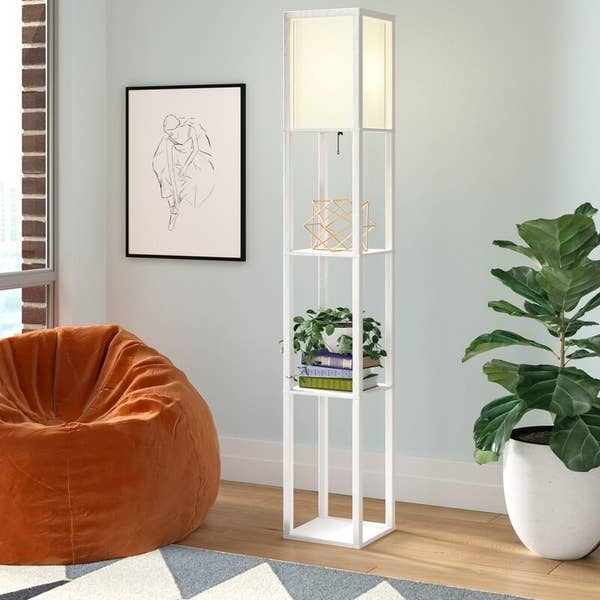 White lamp shelf with green and purple books and a golden geometric display stands beside a green plant in a white pot and an orange bean bag