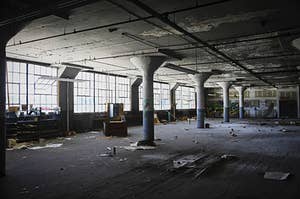 Photograph of the interior of an abandoned textile mill, featuring a wide open room with pillars, wooden floors and remaining debris. The large span of window panes radiate light onto the immensely large wooden floor. 