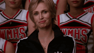 Sue Sylvester from Glee looking amused and covering her mouth in front of a cheerleading squad