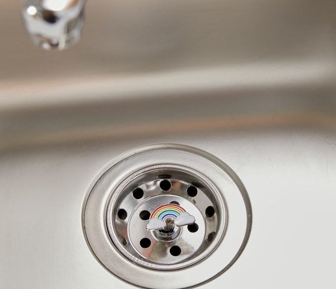 A metal sink strainer with an enamel rainbow charm on top of it 