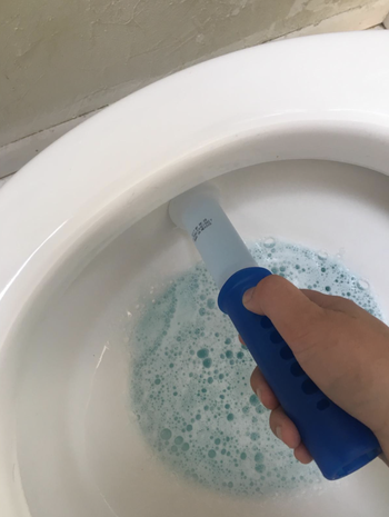 hand holds blue dispenser to place toilet-cleaning stamp on side of toilet bowl