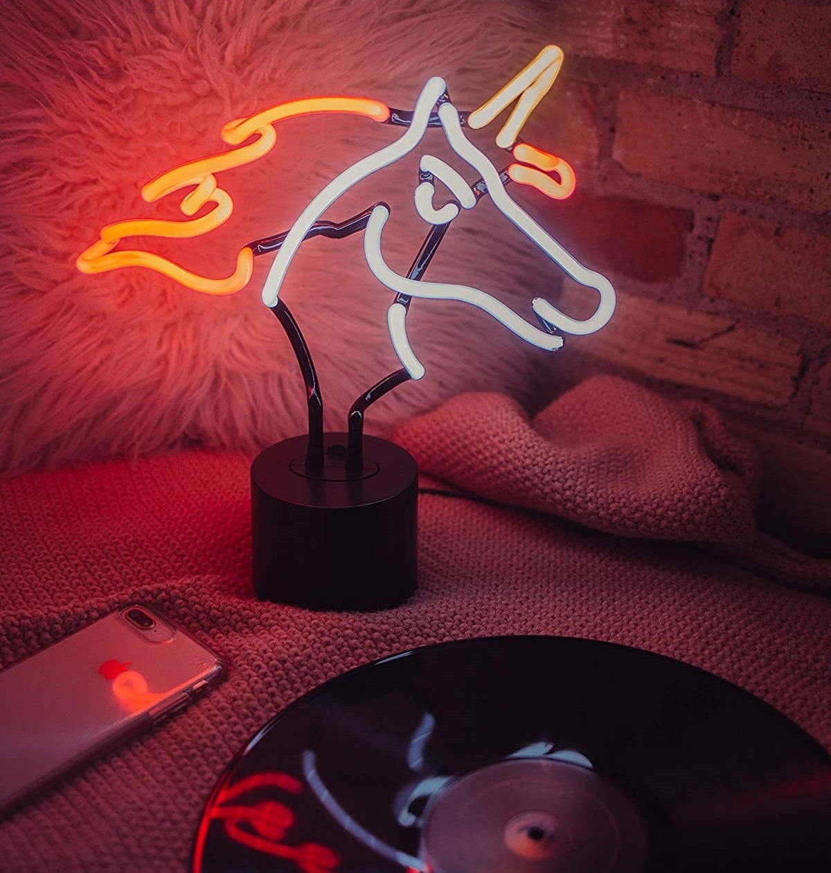 A unicorn-shaped neon light perched on a bed next to a vinyl record, phone, and fluffy pillow