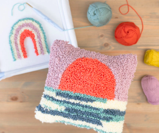 fuzzy pillow that has design of sun setting over sea next to rainbow thread design on a canvas with balls of yarn nearby 