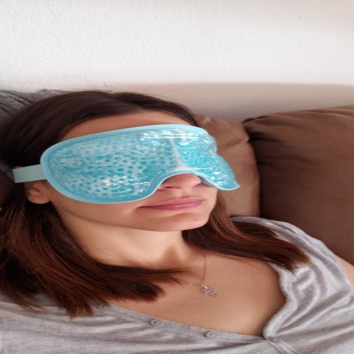Reviewer uses blue cooling eye mask on their forehead while sitting on a couch