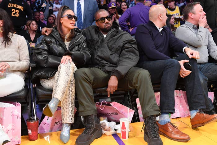 Kim Kardashian and Kanye West sit courtside at a basketball arena, each wearing fashionable outfits and sunglasses
