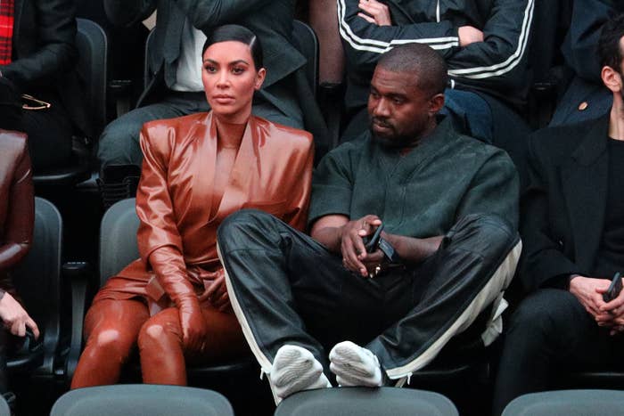 Kardashian wears a fashionable leather bodysuit, sitting next to Kanye, who has his feet up on the chair in front of him