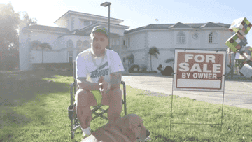 Jake sits in front of his house next to a for sale sign