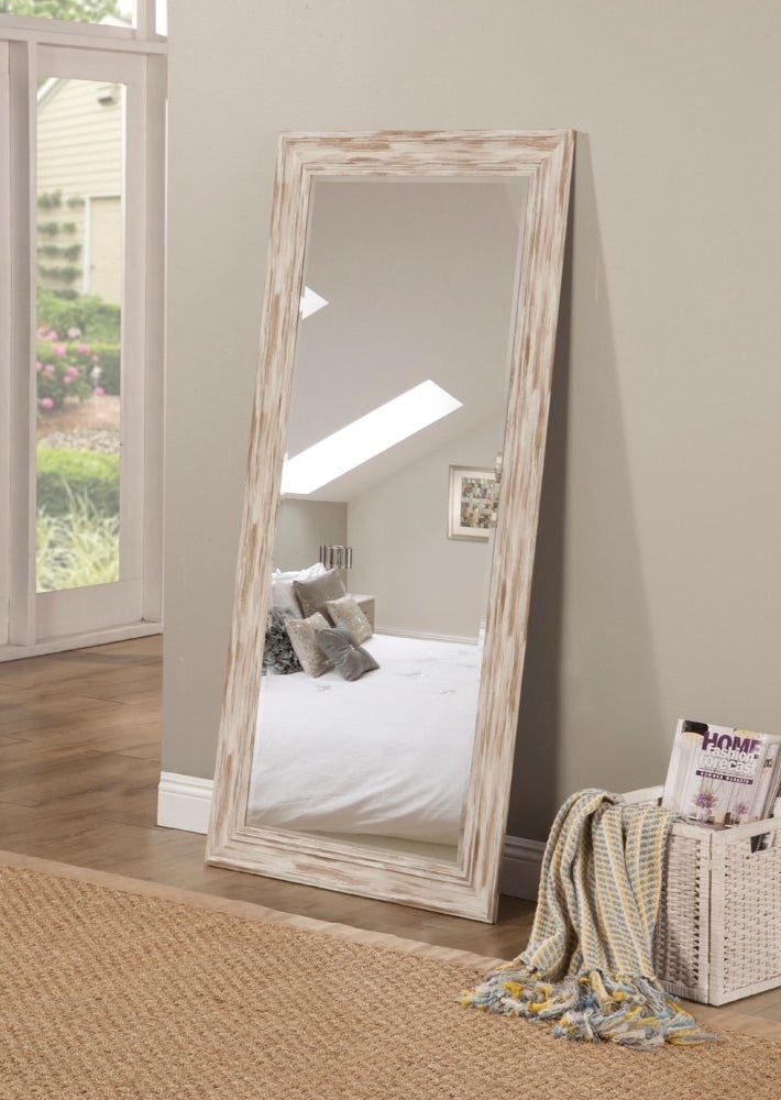 A full-length mirror resting on the floor. The mirror&#x27;s frame is white and brown wood
