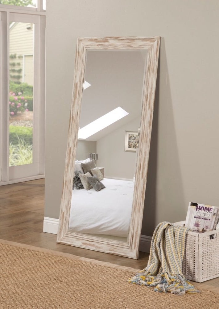 A full-length mirror resting on the floor. The mirror&#x27;s frame is white and brown wood