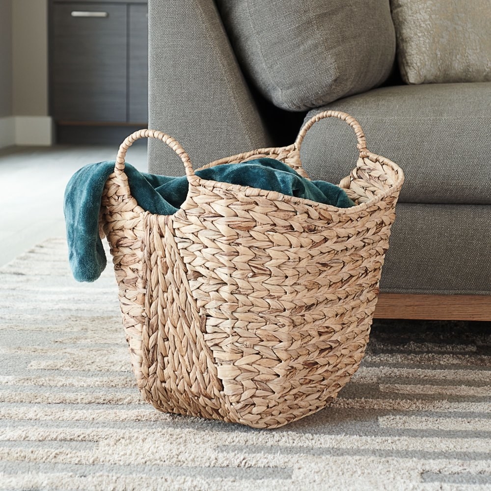 a tan wicker basket with two handles on either side holding a blanket