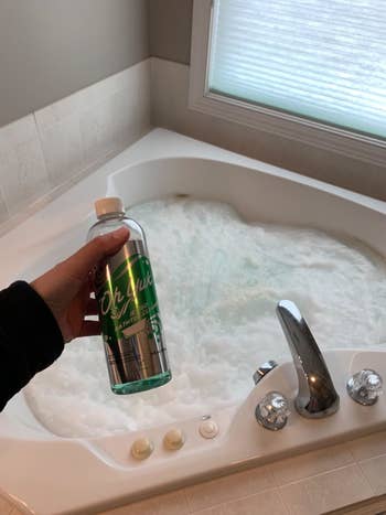 A reviewer holding up the bottle of cleaner in front of their tub which is filled with bubbles