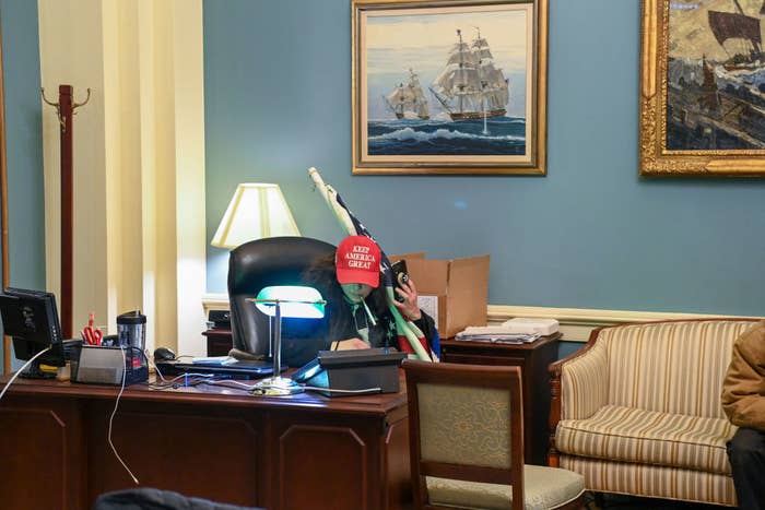 A rioter wearing a 'MAGA' hat and holding an American flag sits at a desk