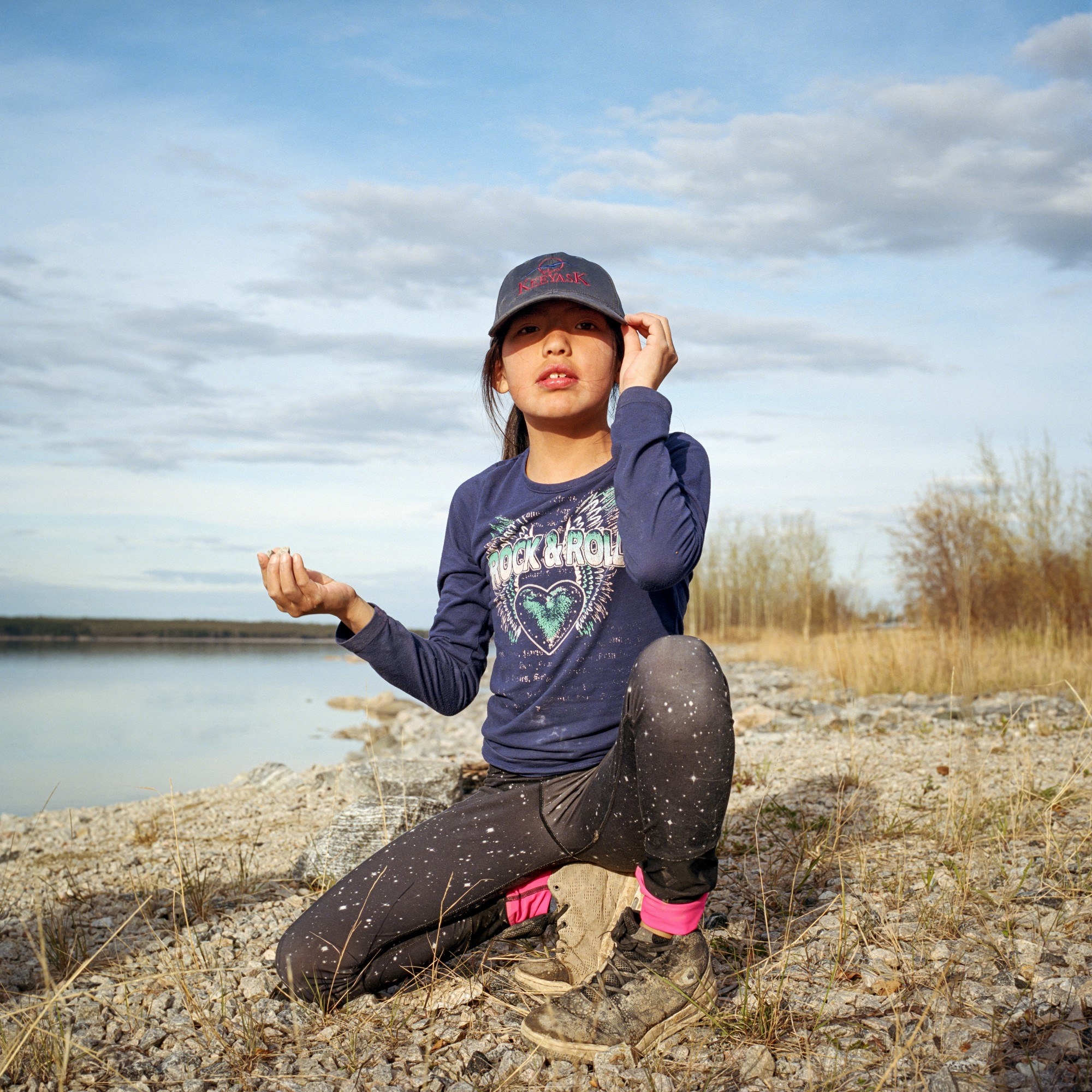 A young girl in a baseball cap kneels by a riverbank in a long-sleeve shirt that says &quot;rock and roll&quot;