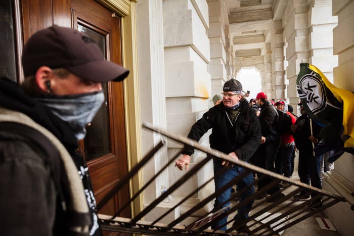 A man carrying a ladder-like object thrusts it into a door as he stands by a crowd of people in a cement-walled hallway