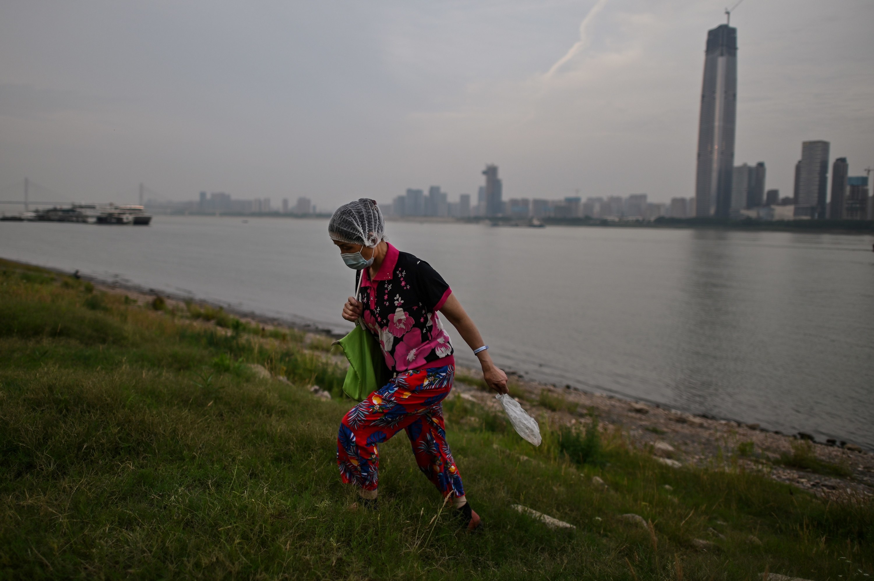A woman wearing a face mask walks on the banks of a river with a city in the background