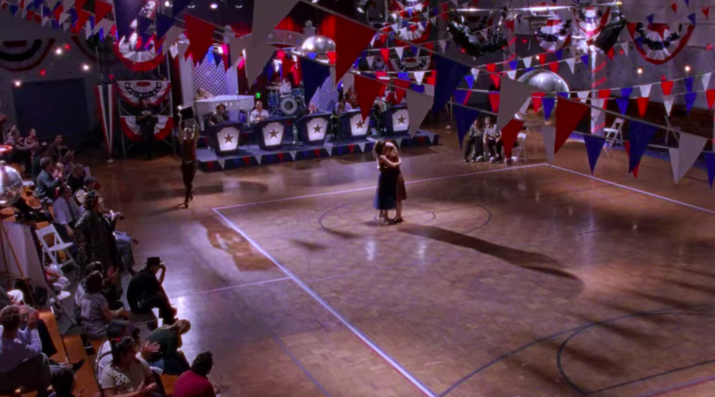 Rory and Lorelai hugging as Kirk dances around the gym with the trophy