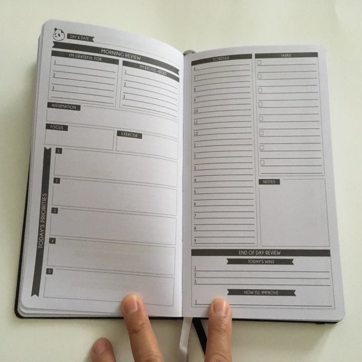 A reviewer photo of a hand holding the planner open to show a daily spread with a morning review and top priorities on the left page, and a schedule, task list, and an end-of-day review on the right page 