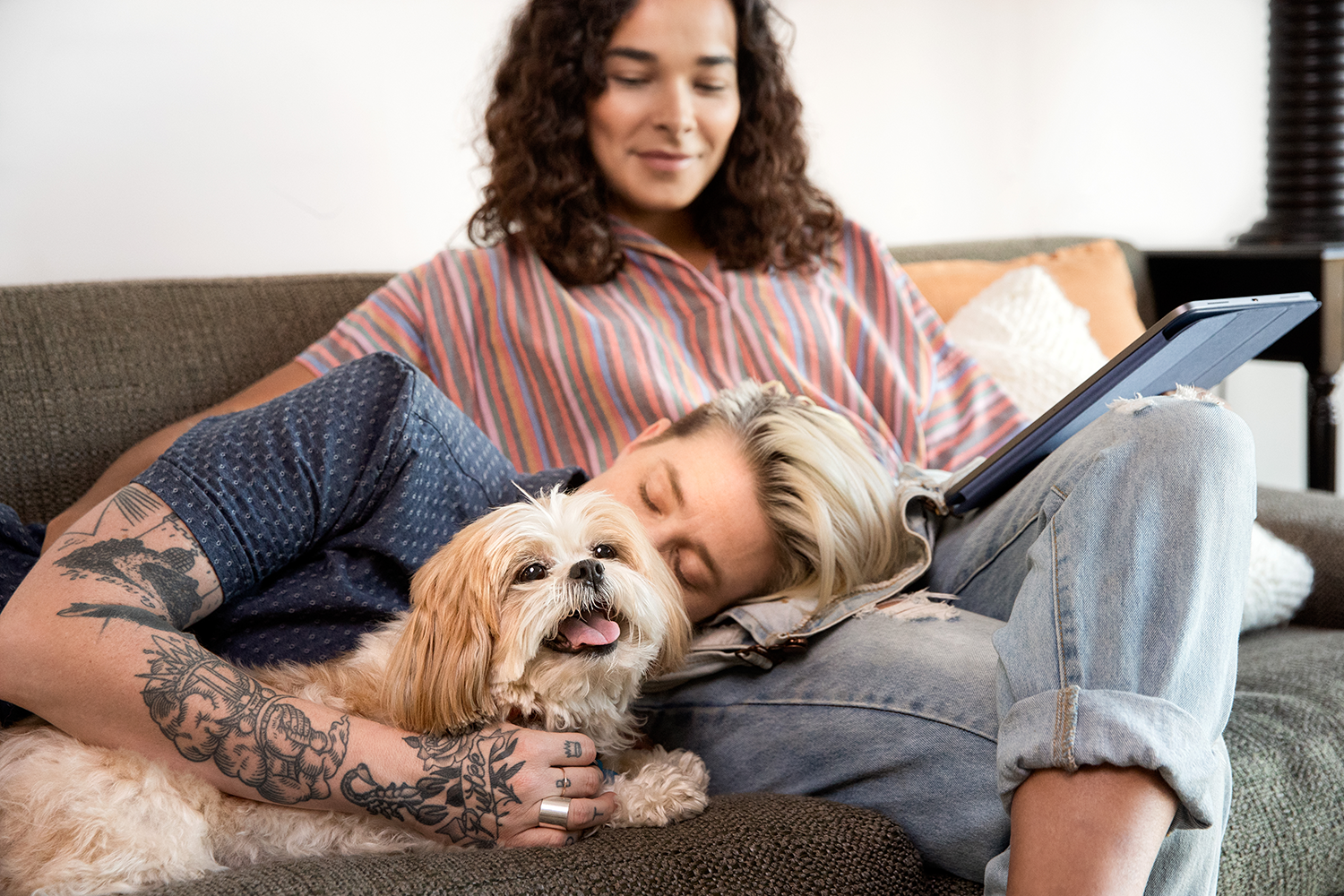 A woman lying with a dog on the couch with her head in the lap of another woman 