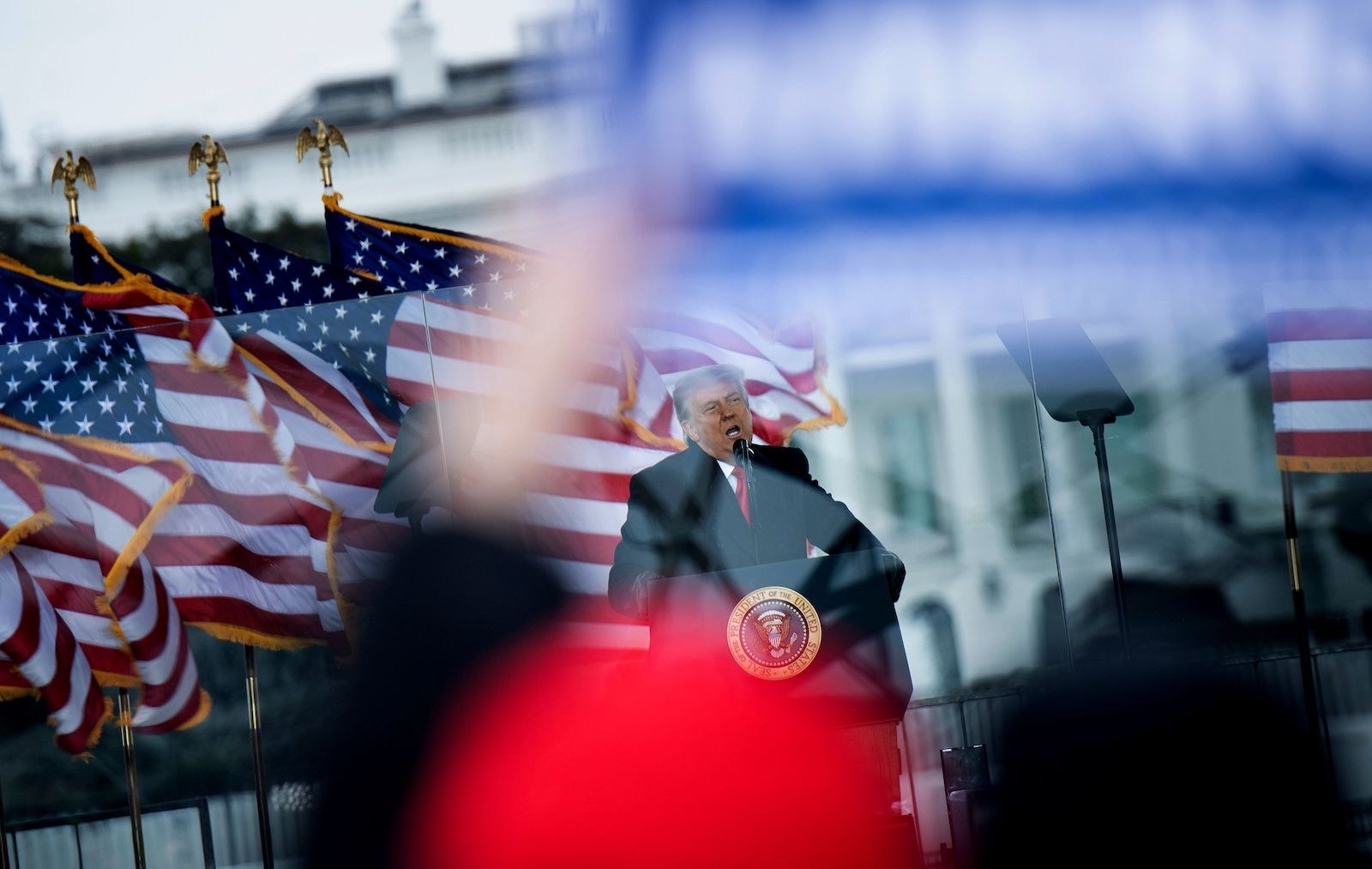 Trump stands behind a lectern with a presidential seal, in front of US flags waving in the wind; in the foreground, out-of-focus supporters wave signs and flags