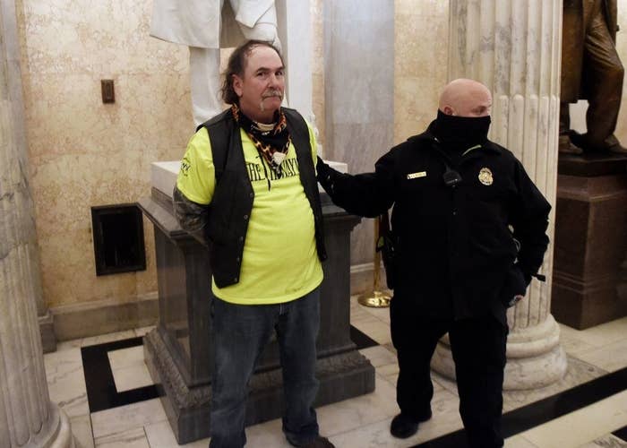 A man stands in the Capitol in front of a statue with his hands behind his back as a police officer stands next to him