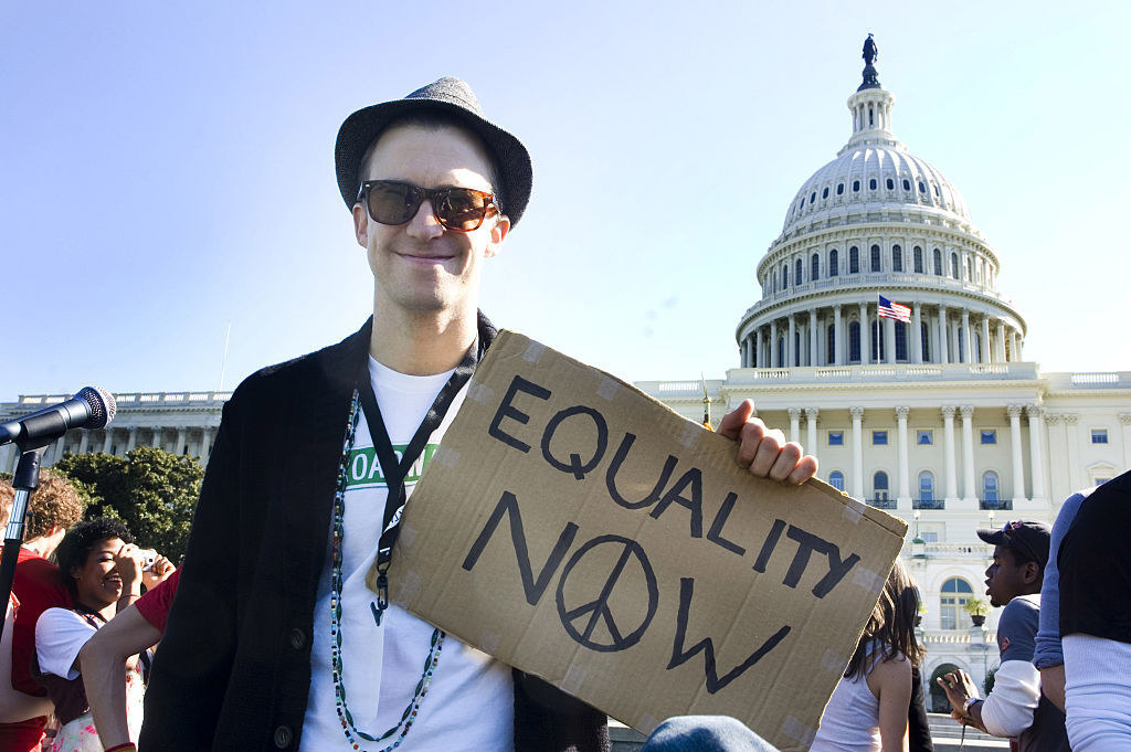 A man standing outside the Capitol, smiling at the camera and holding a sign that says, "Equality now"