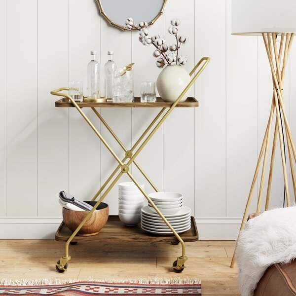 The brass and wood bar cart which has an X design, two shelves, and four wheels