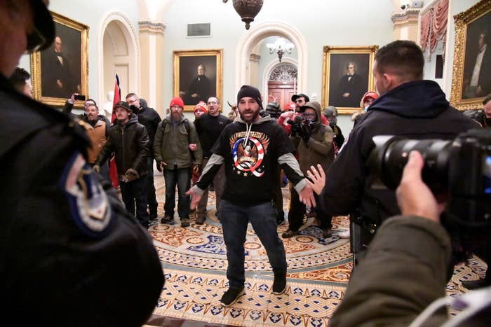 A man wearing a black T-shirt with &quot;Q&quot; and an eagle on it stands in front of a group of maskless men, facing police