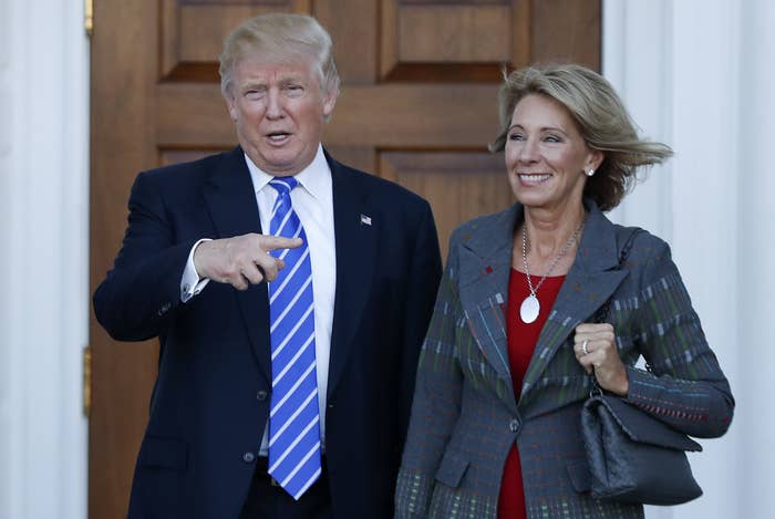 A smiling Trump points to a smiling DeVos as they stand together outside the White House on a sunny day