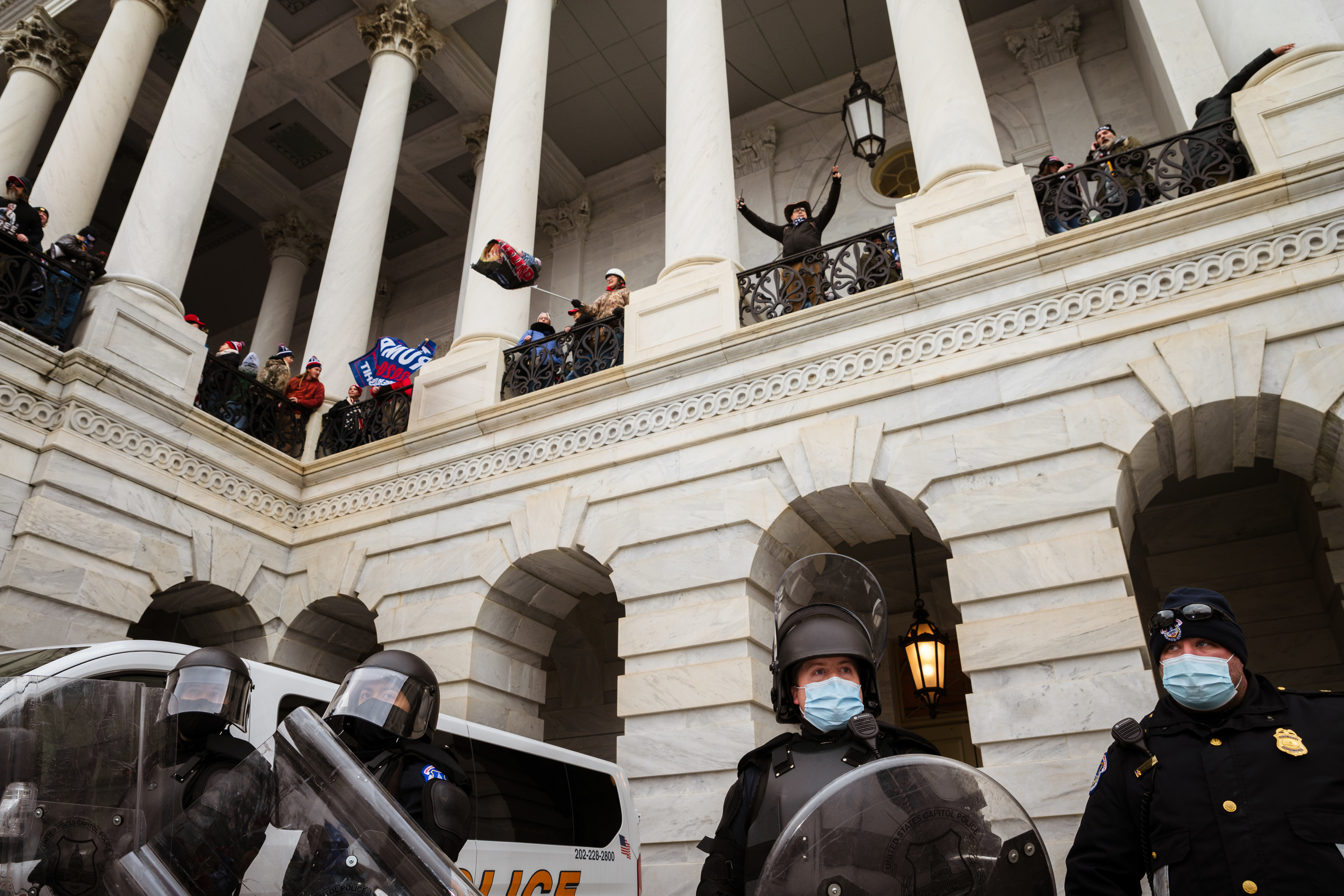 Trump supporters wave flags between columns at the Capitol building on a second floor; below them, police wear face masks, helmets, and shields