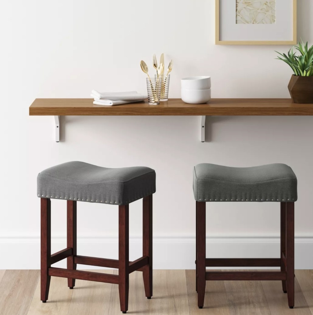 A pair of bar stools with gray cushions, nailhead detail, and wooden legs