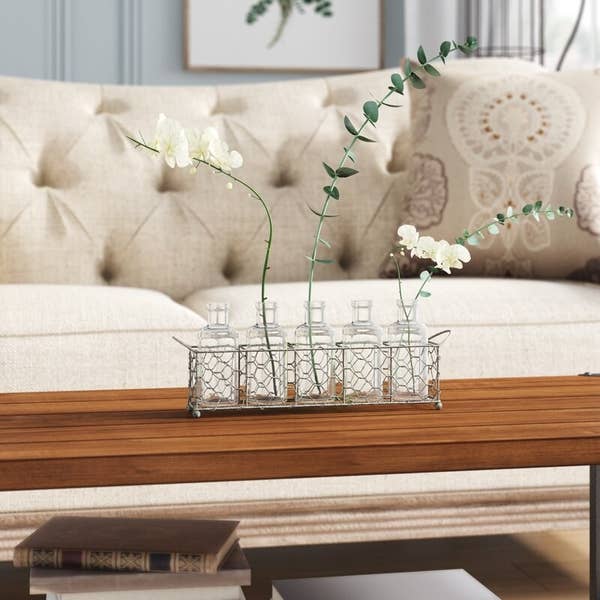 Distressed wire tray holds six glass bottles and white and green wildflowers on a brown table near a tan sofa