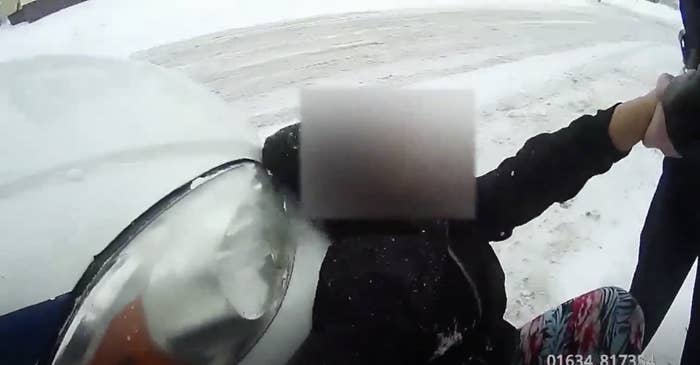 A child with a blurred-out face sits on the snow-covered ground against a police car