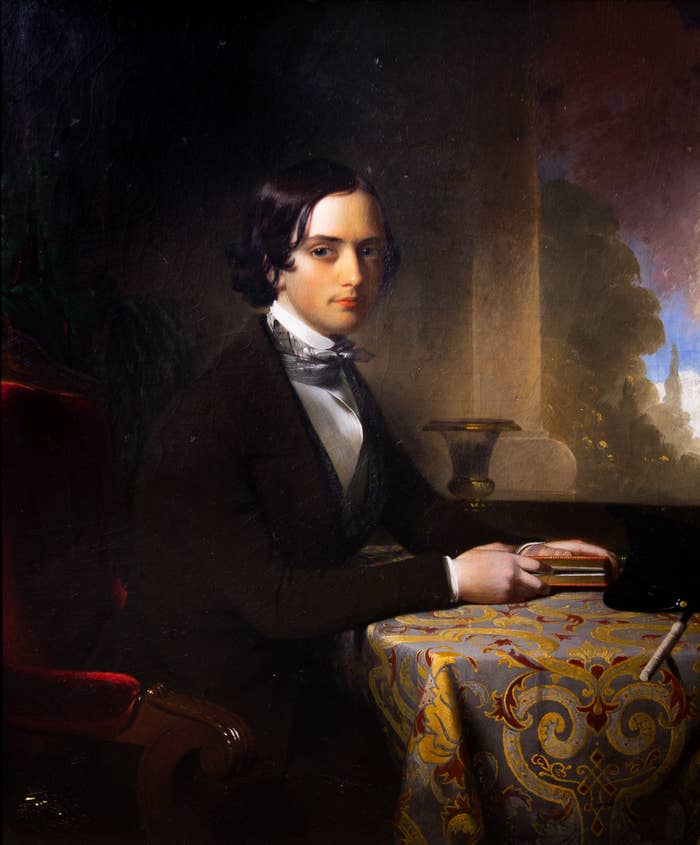 A portrait of a young man sitting at a table facing the right side of the painting with his head turned over his right shoulder towards the viewer. 