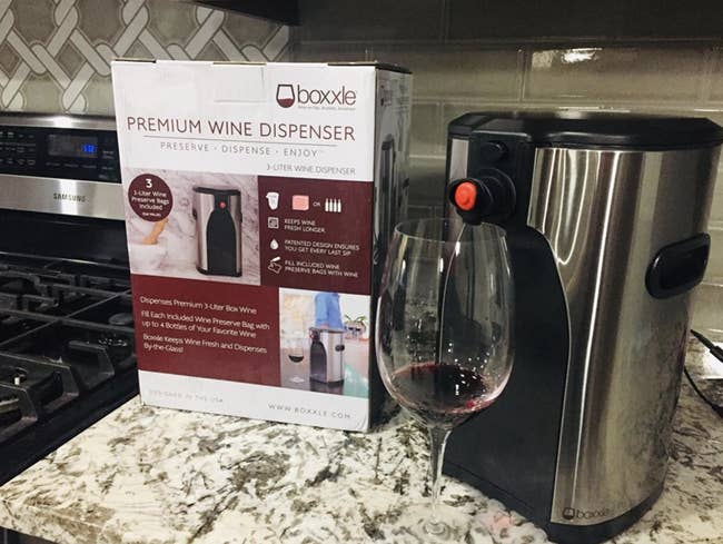 reviewer pic of the silver sleek wine dispenser on a counter, with a wine glass in front of it, pouring red wine into it