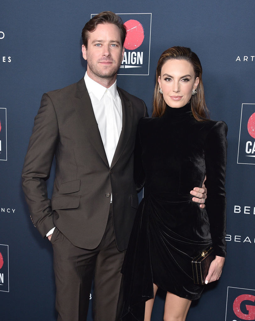 Armie, wearing a suit, and Elizabeth Chambers, wearing a short velvet dress and carrying a clutch, pose on a red carpet