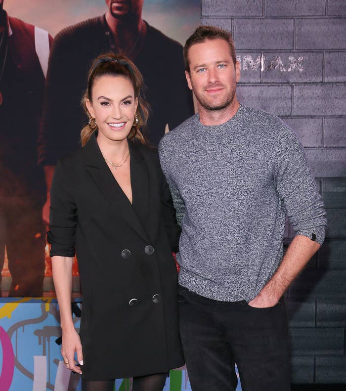 Elizabeth Chambers, wearing a blazer dress and pantyhose, and Armie smile as they pose for photos on a red carpet