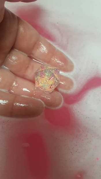 hand holds glittery 20 sided dice in tub with pink water