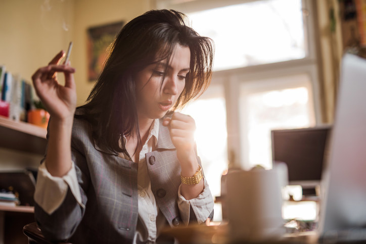 A woman holding a cigarette and coughing as she sits at her desk