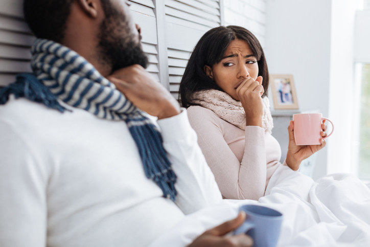 A man, who&#x27;s holding a coffee mug, and a woman, who&#x27;s coughing and also. holding a mug, sitting in bed