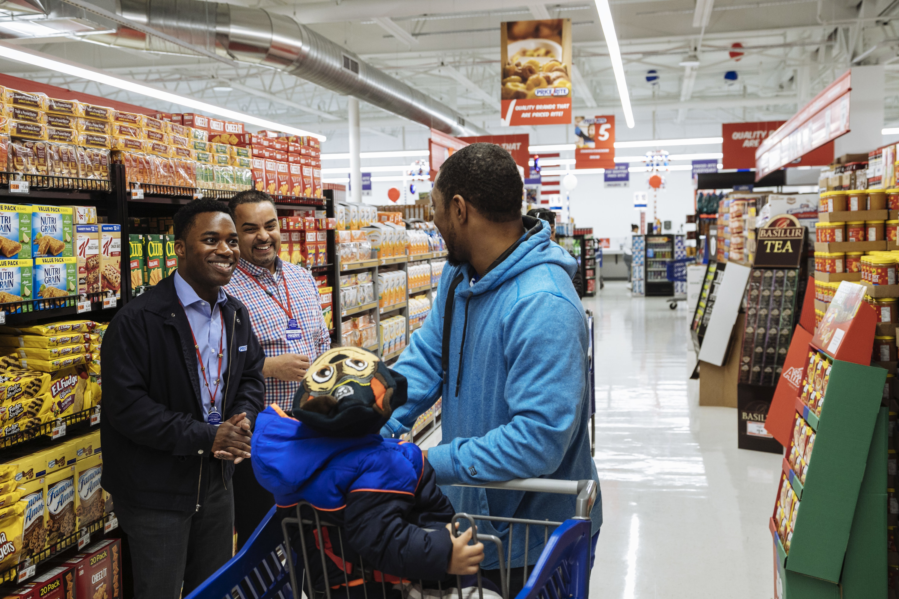 A man in a hoodie pushing a shopping cart talks to two Price Rite employees in the cookie aisle of a supermarket