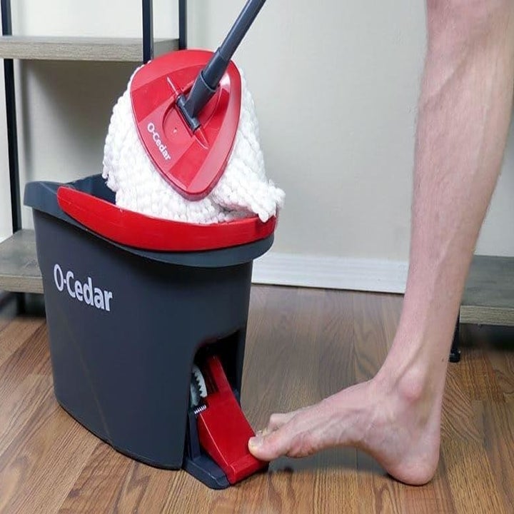A reviewer photo of the mop bucket and mop with a foot pressing down on a pedal that powers the built-in wringer 