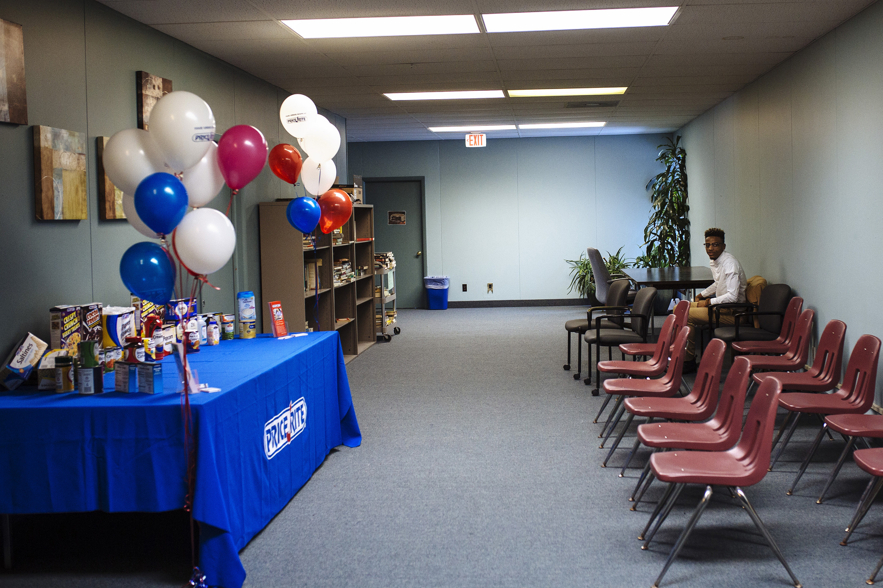 A man sits alone in a room with empty chairs and a table with snacks and balloons