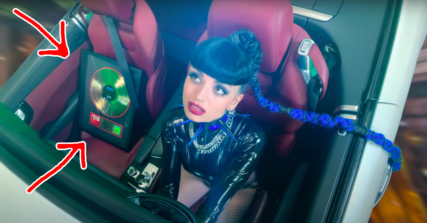 Arrows pointing at the plaque in the passenger seat as Rebecca, dressed in a leather bodysuit, drives with her long braided ponytail whipping in the wind
