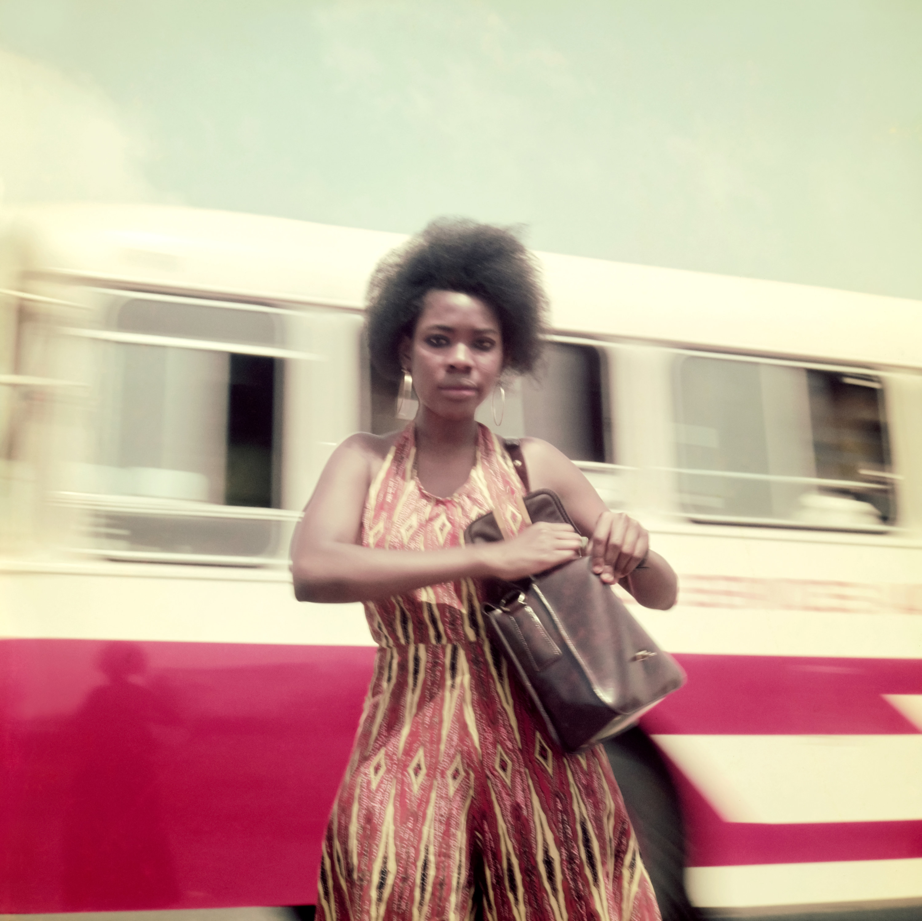 A woman in a dress and purse in front of a moving bus