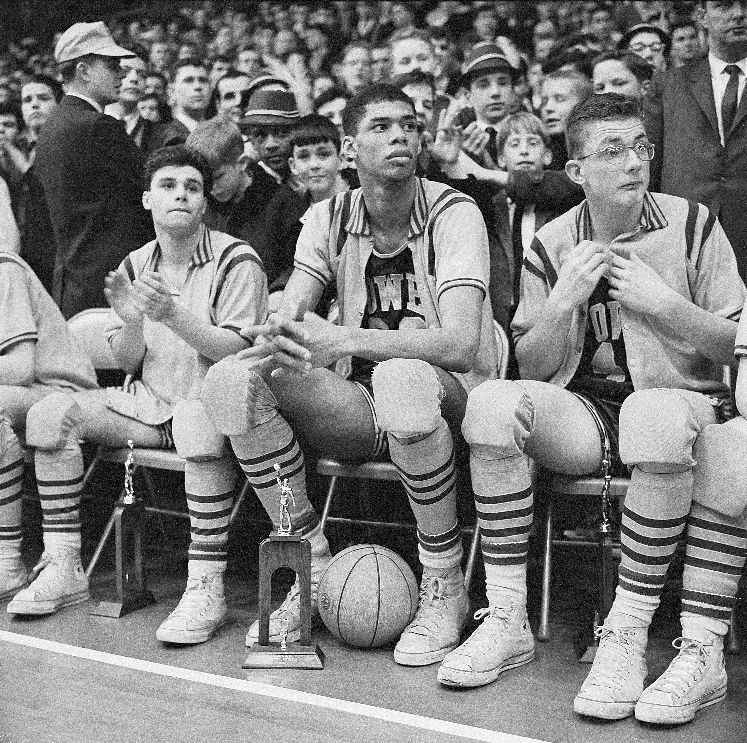 A young Kareem Abdul-Jabbar sitting on the sidelines of a basketball game with a trophy