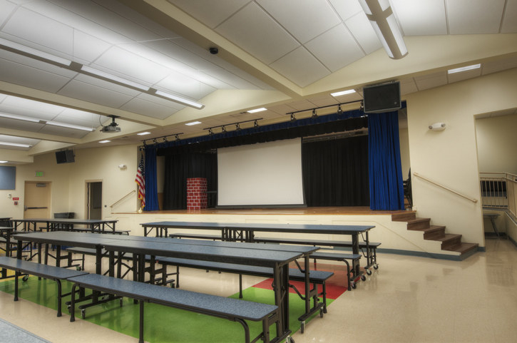 A large, empty school cafeteria with long tables and benches. A stage with a wooden floor, blue curtains, and a white screen is in the background. An American flag is visible