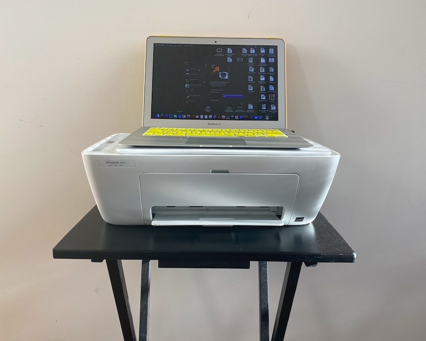 A laptop sits on top of a printer, which is placed on a TV tray.