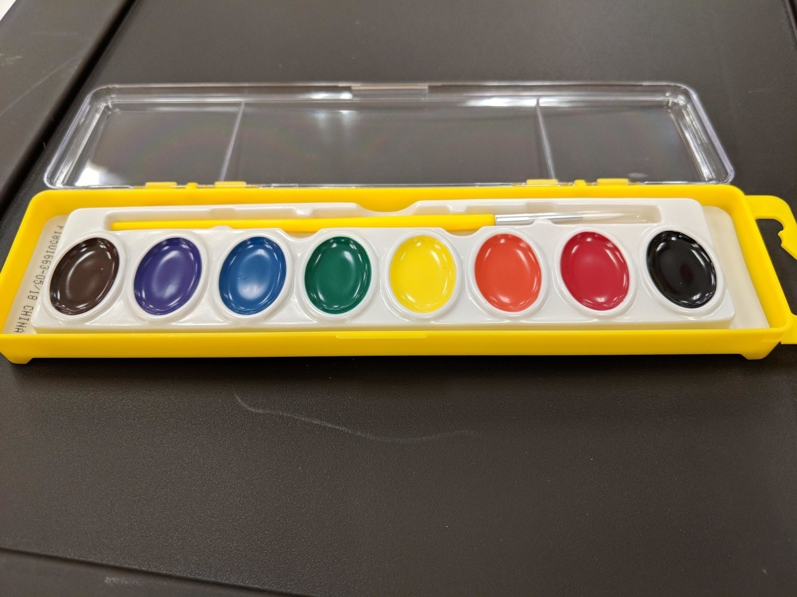 An open watercolor paint set with eight different colors and a paintbrush inside a yellow plastic case